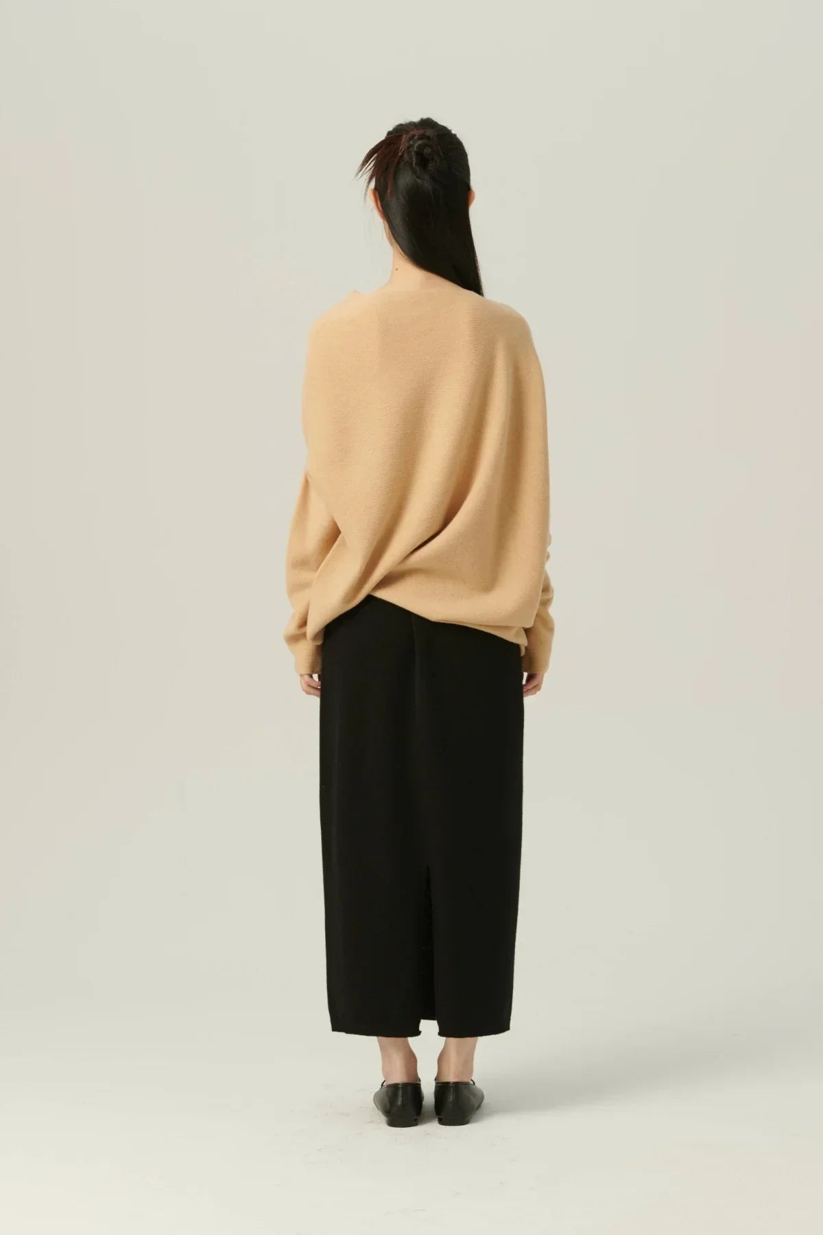 Woman wearing beige oversized knit sweater and black midi skirt with back slit, standing against plain background