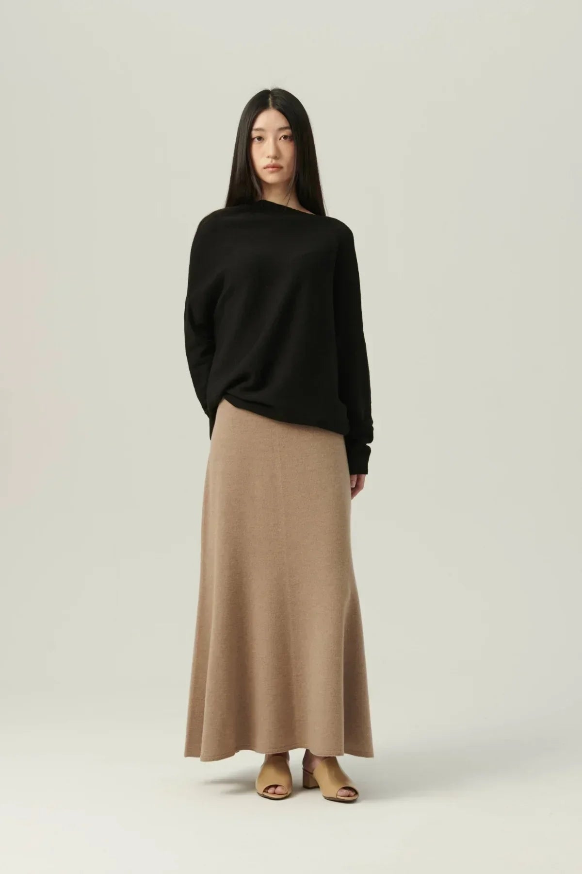 Woman wearing black off-shoulder sweater, beige maxi skirt, and tan open-toe heels on plain background