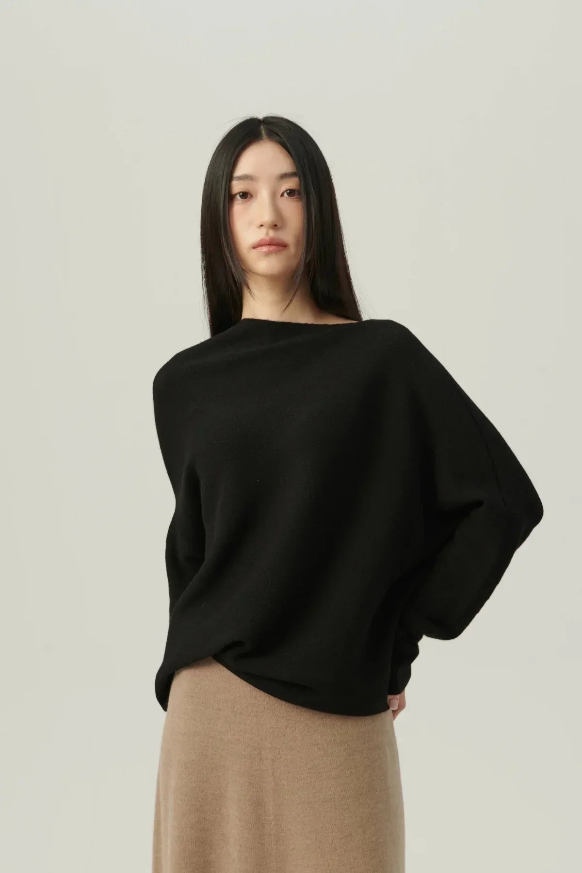 Woman wearing black oversized knit sweater with one-shoulder design and beige skirt on light background