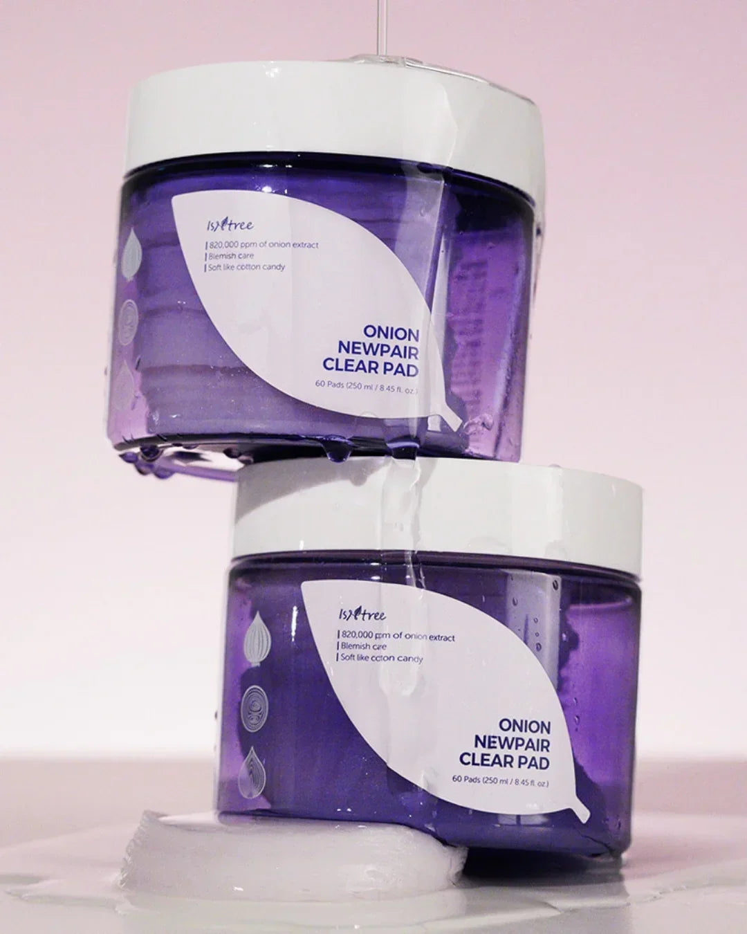 Two stacked purple containers of Isntree Onion Newpair Clear Pad with water droplets on a cotton pad against a soft pink background