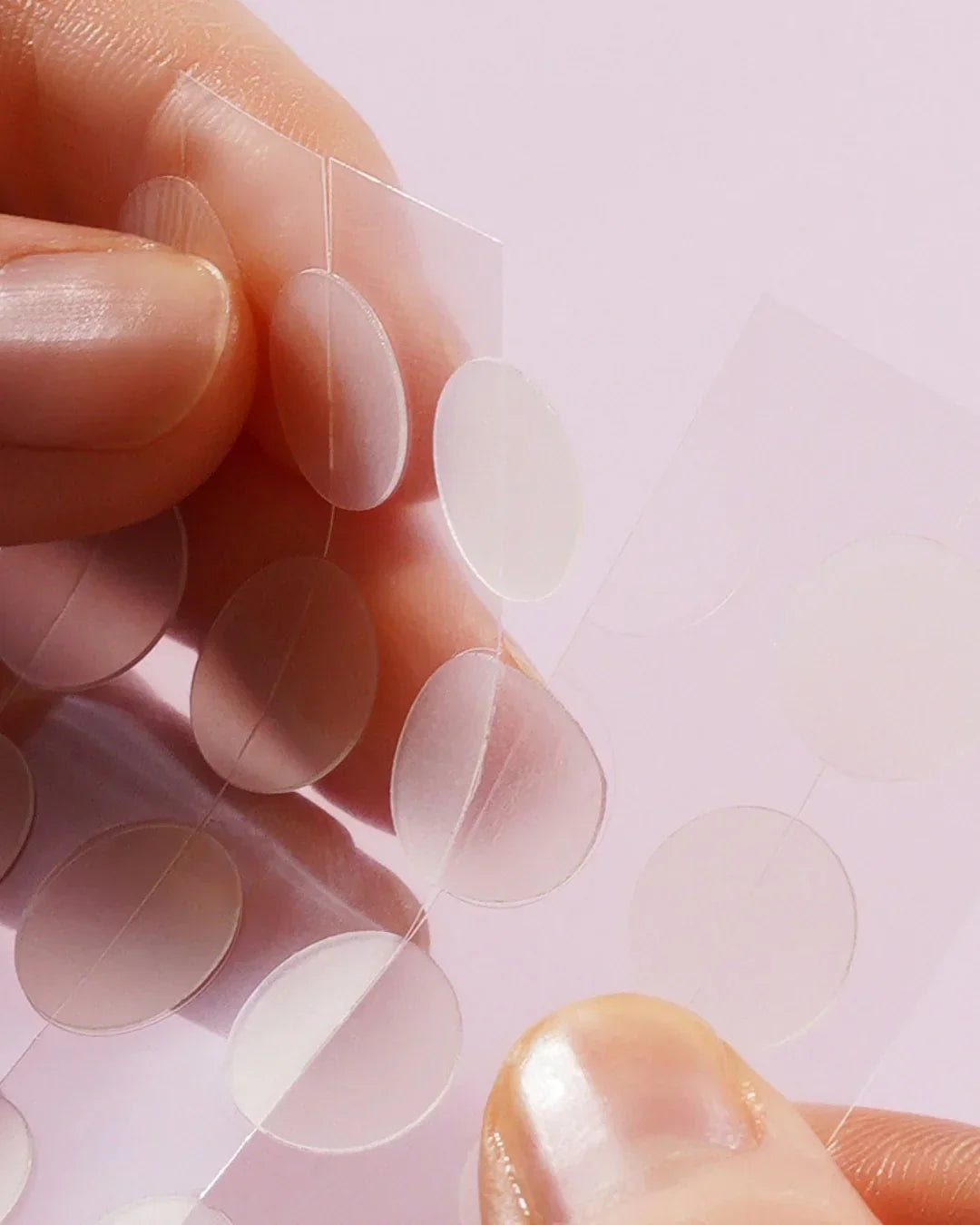 Close-up of hands holding clear acne spot patches with round translucent shapes on light pink background