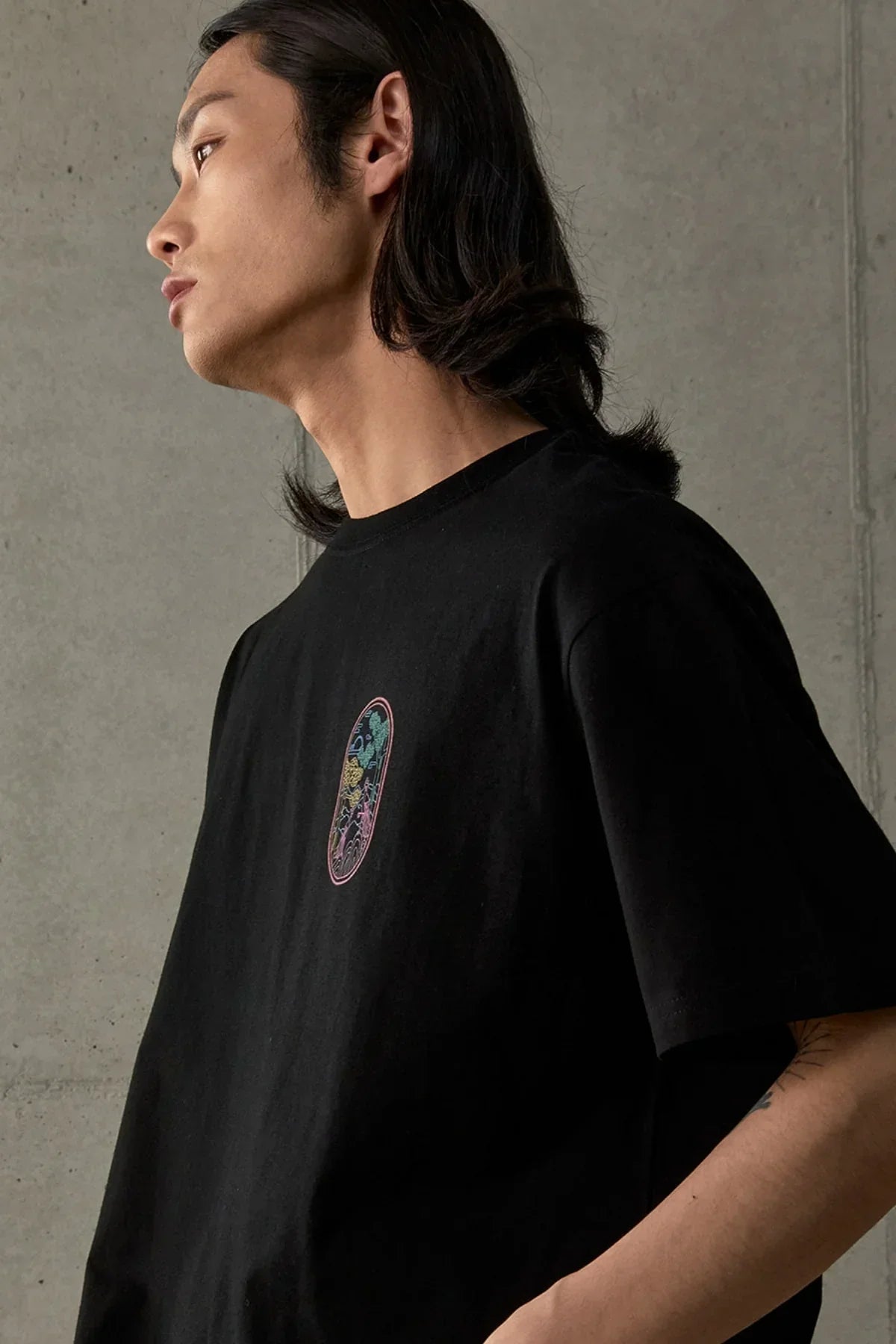 Side view of model wearing black t-shirt with colorful phoenix embroidery on chest against concrete wall