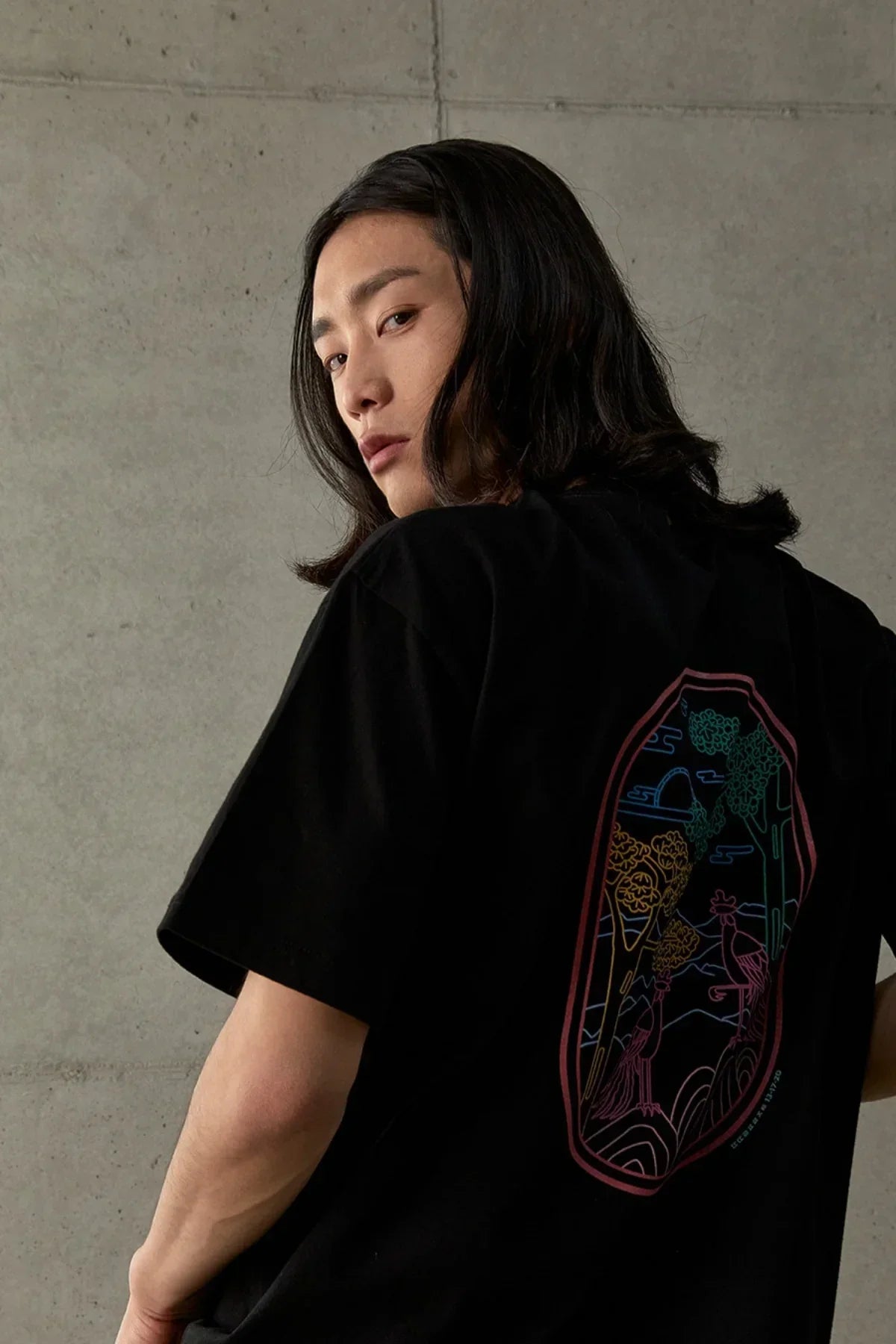 Person wearing black T-shirt with colorful phoenix and tree artwork on back, gray concrete background