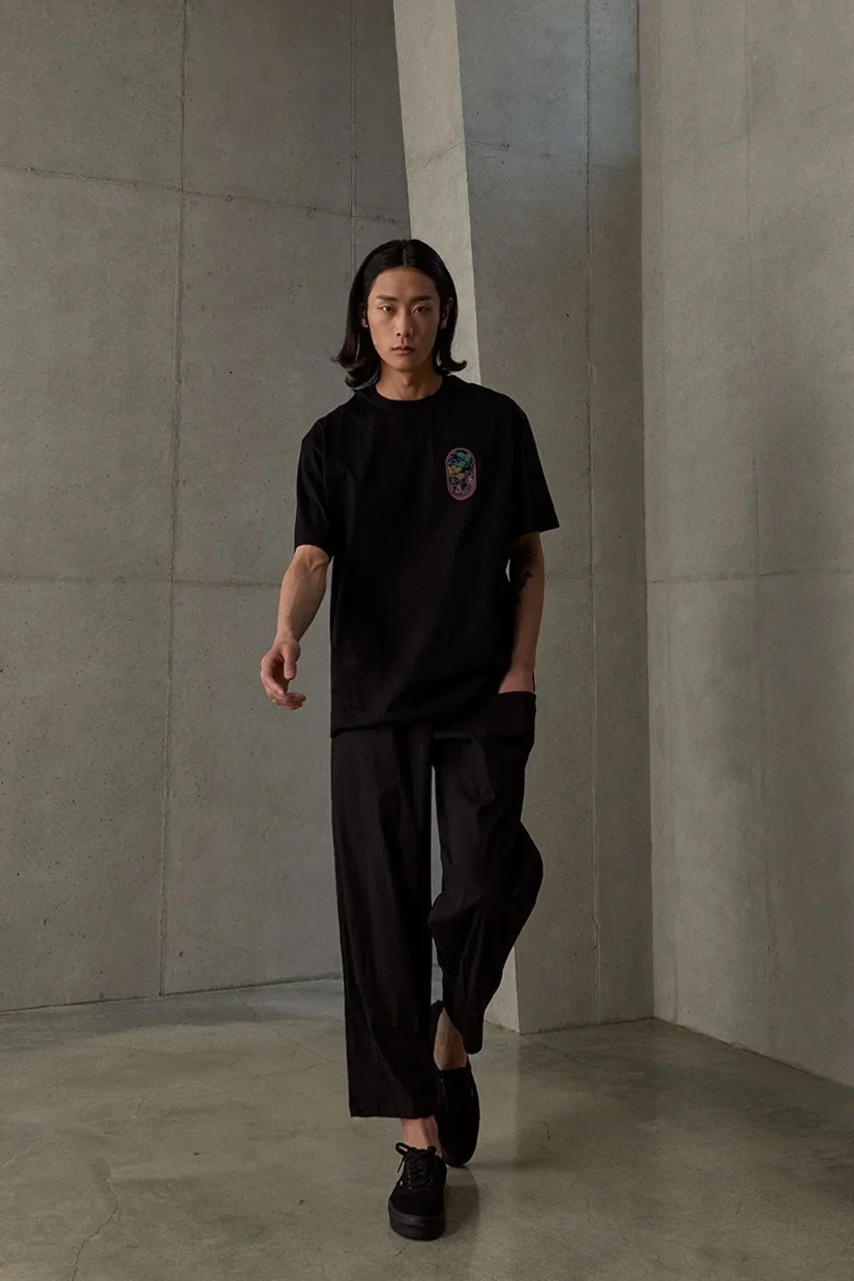 Man wearing black T-shirt with colorful phoenix graphic, black wide-leg pants, and black shoes in minimal concrete room