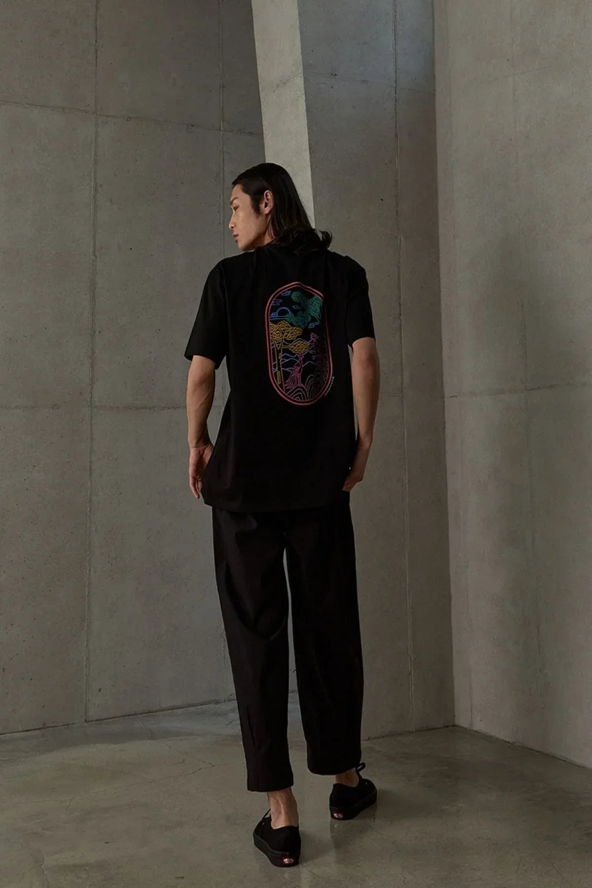 Man wearing black T-shirt with colorful phoenix and tree graphic on back in minimalist concrete room