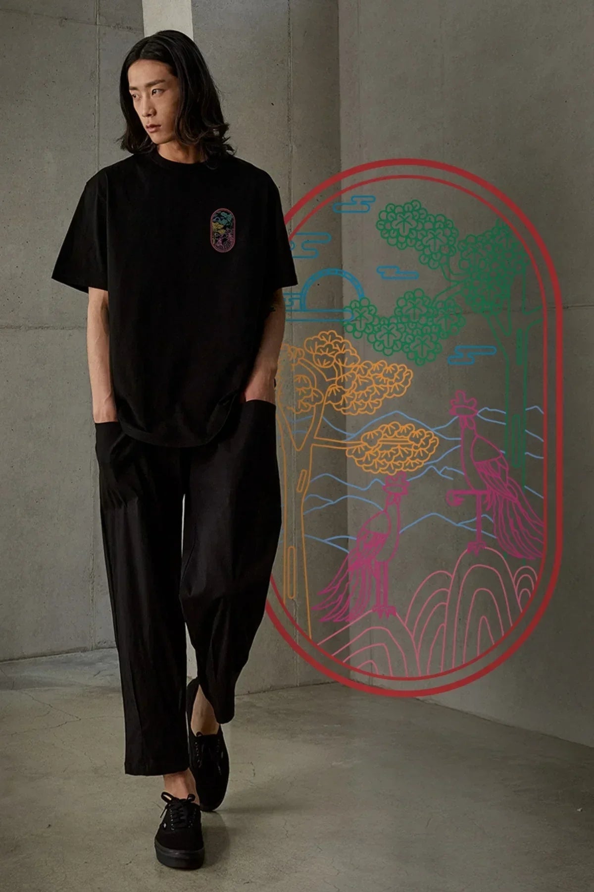 Man wearing black t-shirt with colorful phoenix graphic, black pants, and black shoes in minimal concrete setting
