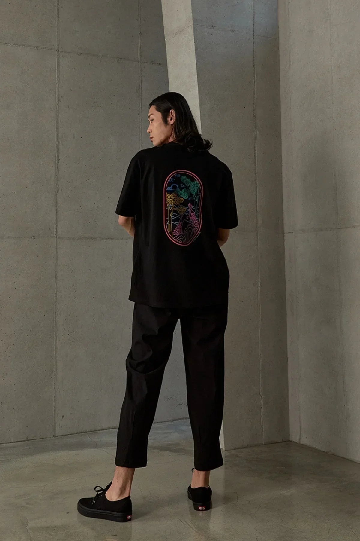 Man in black pants and black T-shirt with colorful abstract phoenix design on back in minimalist concrete room
