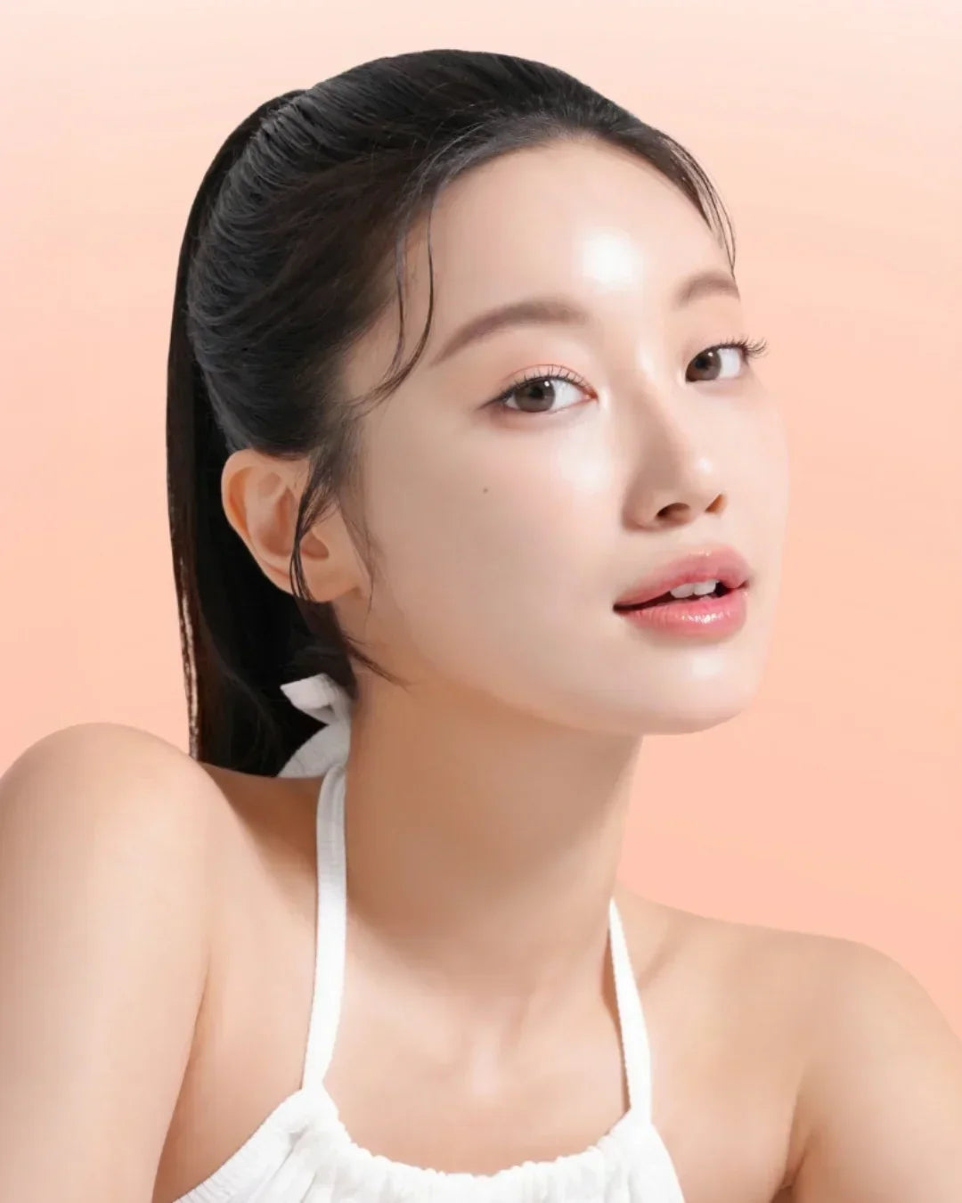 Close-up of woman with glowing skin and white halter top against peach background, skincare beauty