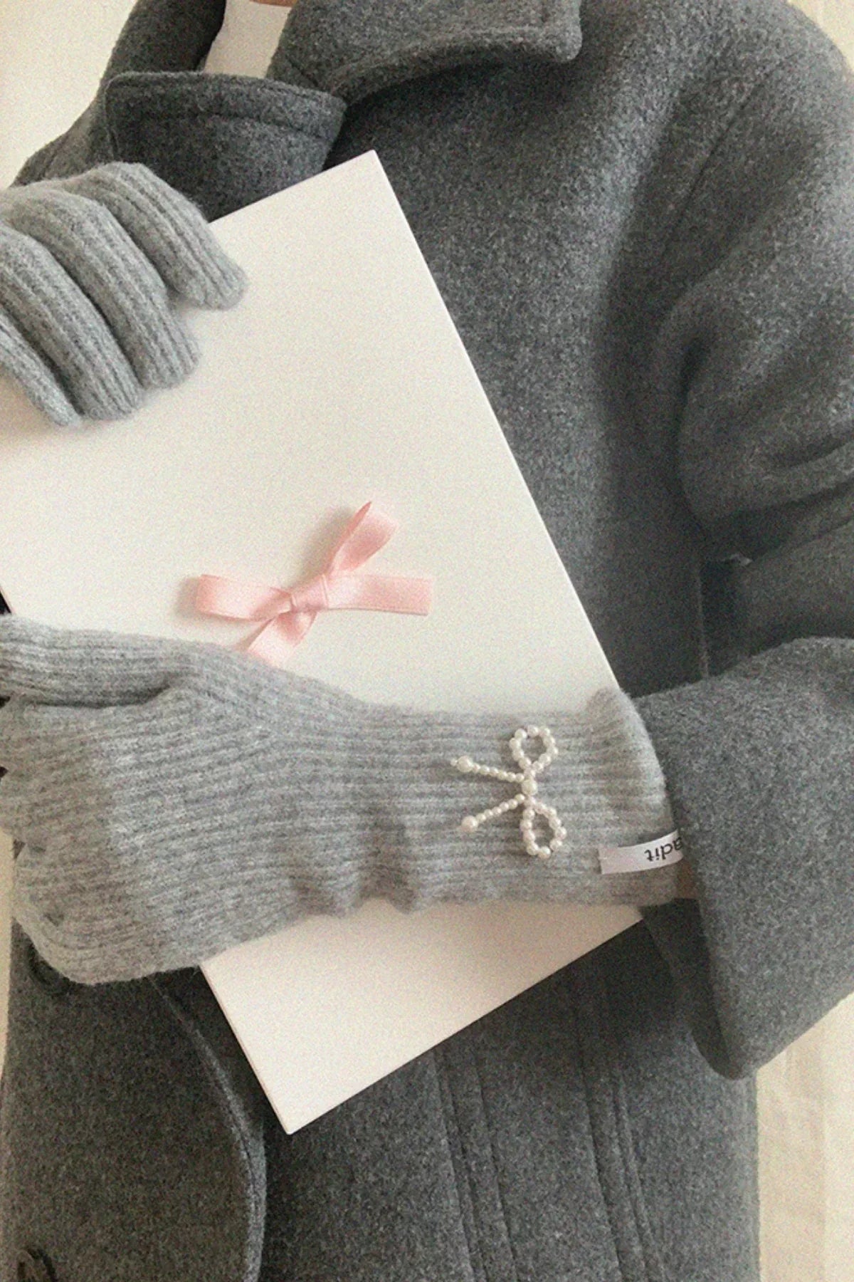Gray ribbed knit gloves with pearl ribbon detail holding white gift box with pink bow