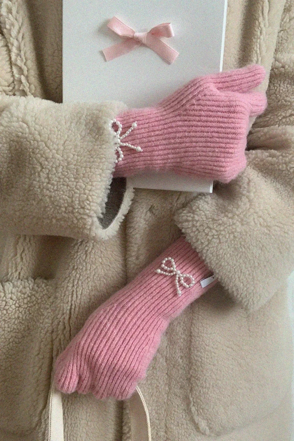 Soft pink ribbed knit gloves with pearl bow detail worn over beige fuzzy coat holding white box and bag