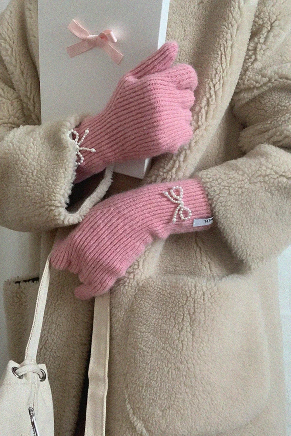 Person wearing pink knit gloves with pearl bow accents holding a white box, beige fleece coat, and white bag