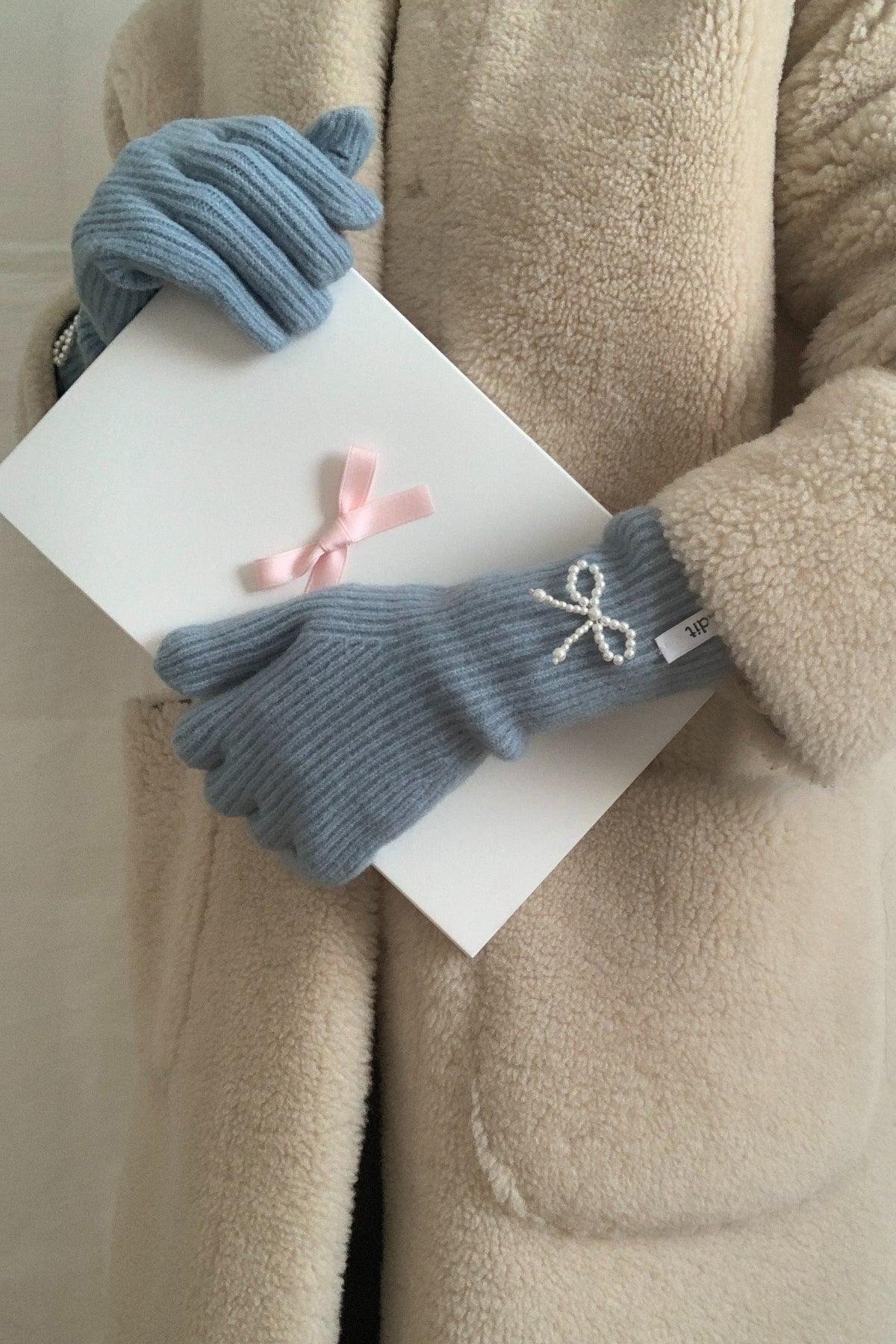 Person wearing cozy sky blue ribbed gloves with pearl bow detail holding a white gift box with pink ribbon