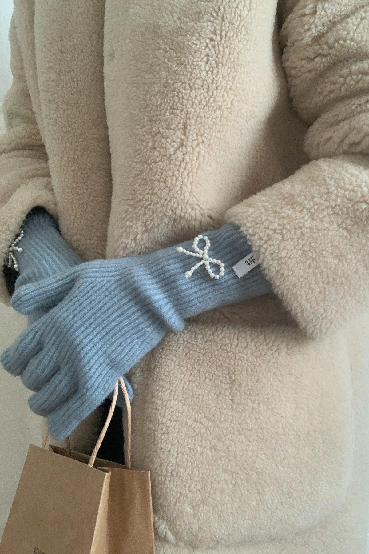 Person wearing sky blue ribbed gloves with pearl bow detail and beige teddy coat holding brown paper bag