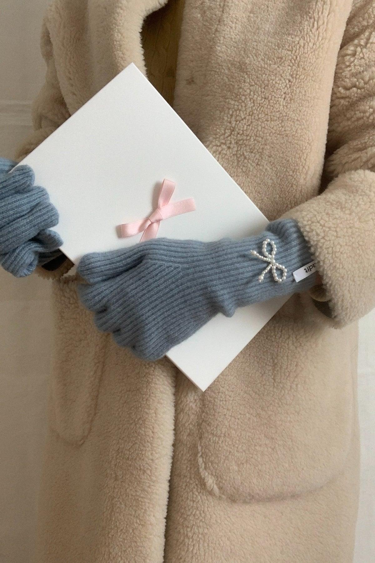 Person wearing sky blue knit gloves with pearl bow detail holding white box with pink ribbon, beige fleece coat