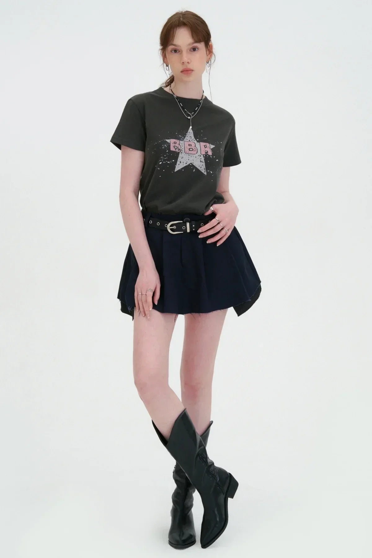 Female model wearing charcoal graphic t-shirt with star design, black pleated mini skirt, and black boots