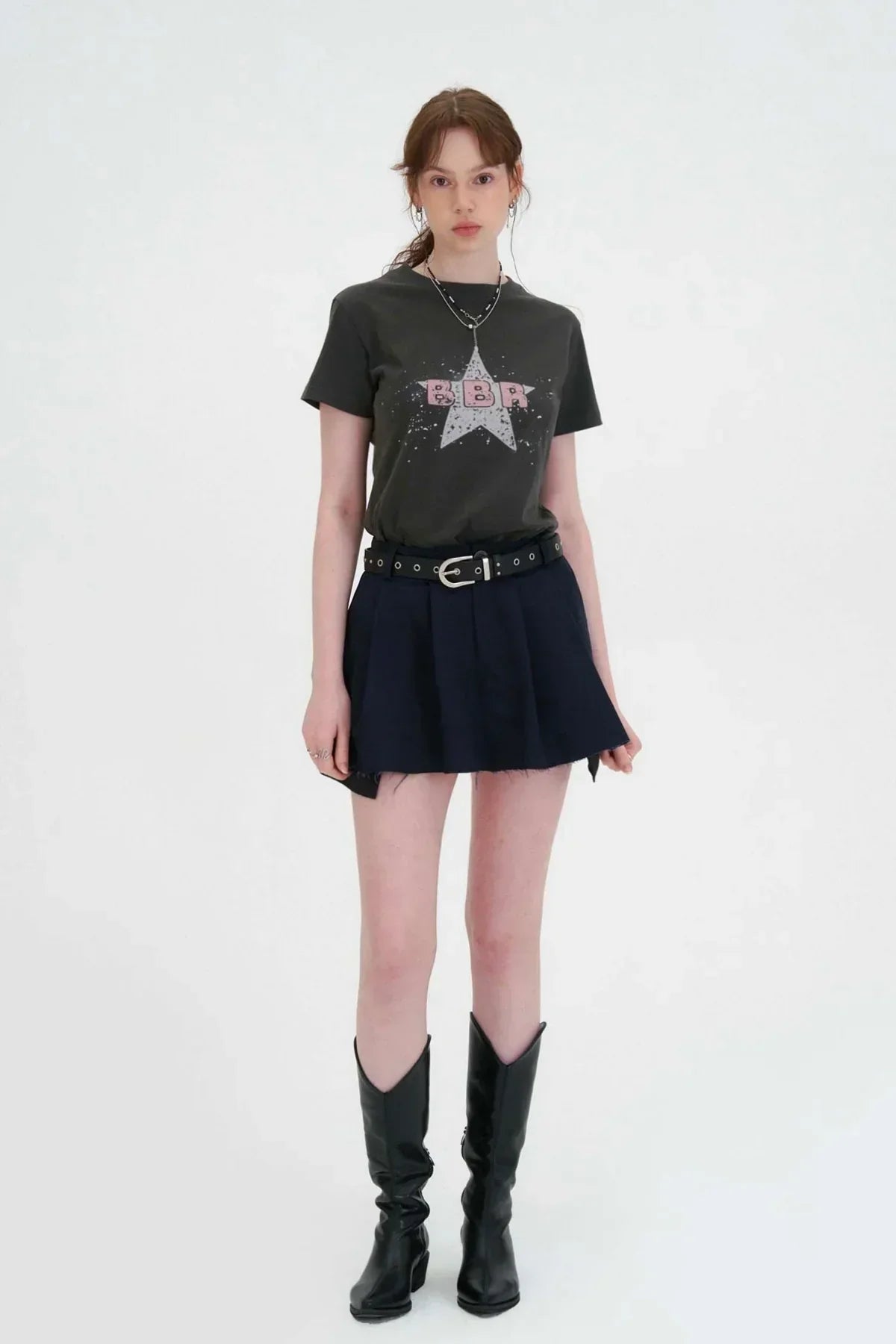 Young woman modeling charcoal graphic t-shirt with star print, pleated black mini skirt, and black knee-high boots