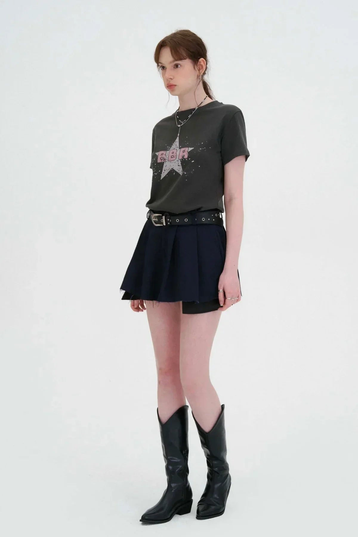 Young woman wearing charcoal graphic t-shirt, black pleated mini skirt, black belt, and black cowboy boots on white background