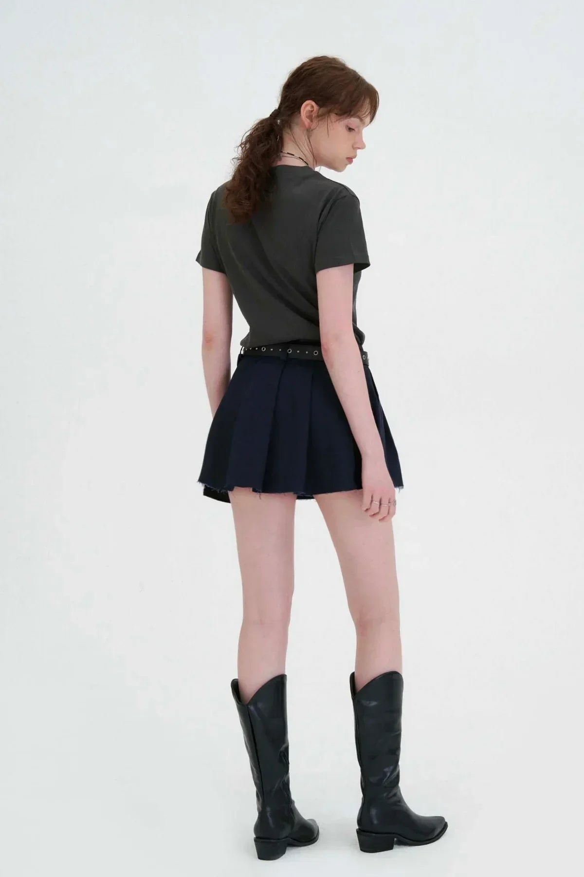 Female model wearing charcoal crop t-shirt, navy pleated skirt, and black cowboy boots on white background