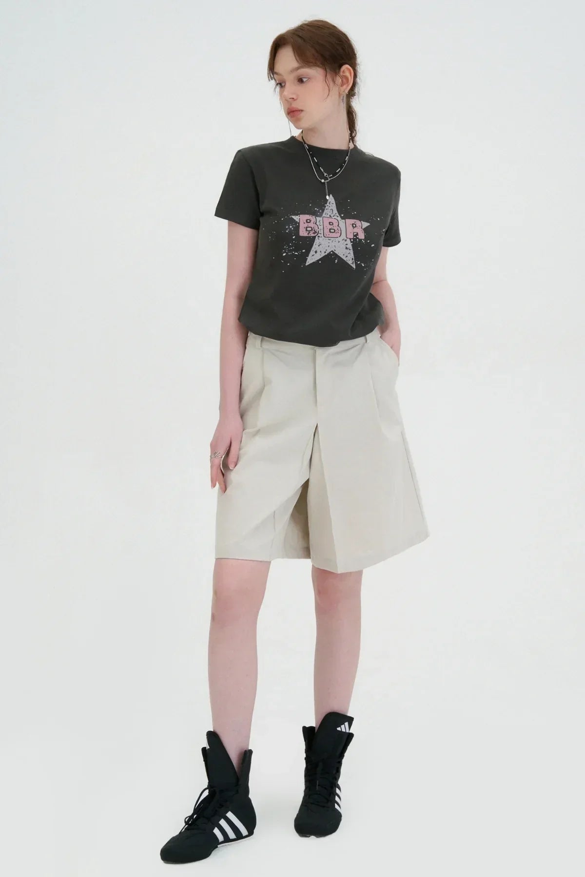 Woman wearing charcoal BBR star graphic t-shirt with beige pleated shorts and black Adidas high-top sneakers
