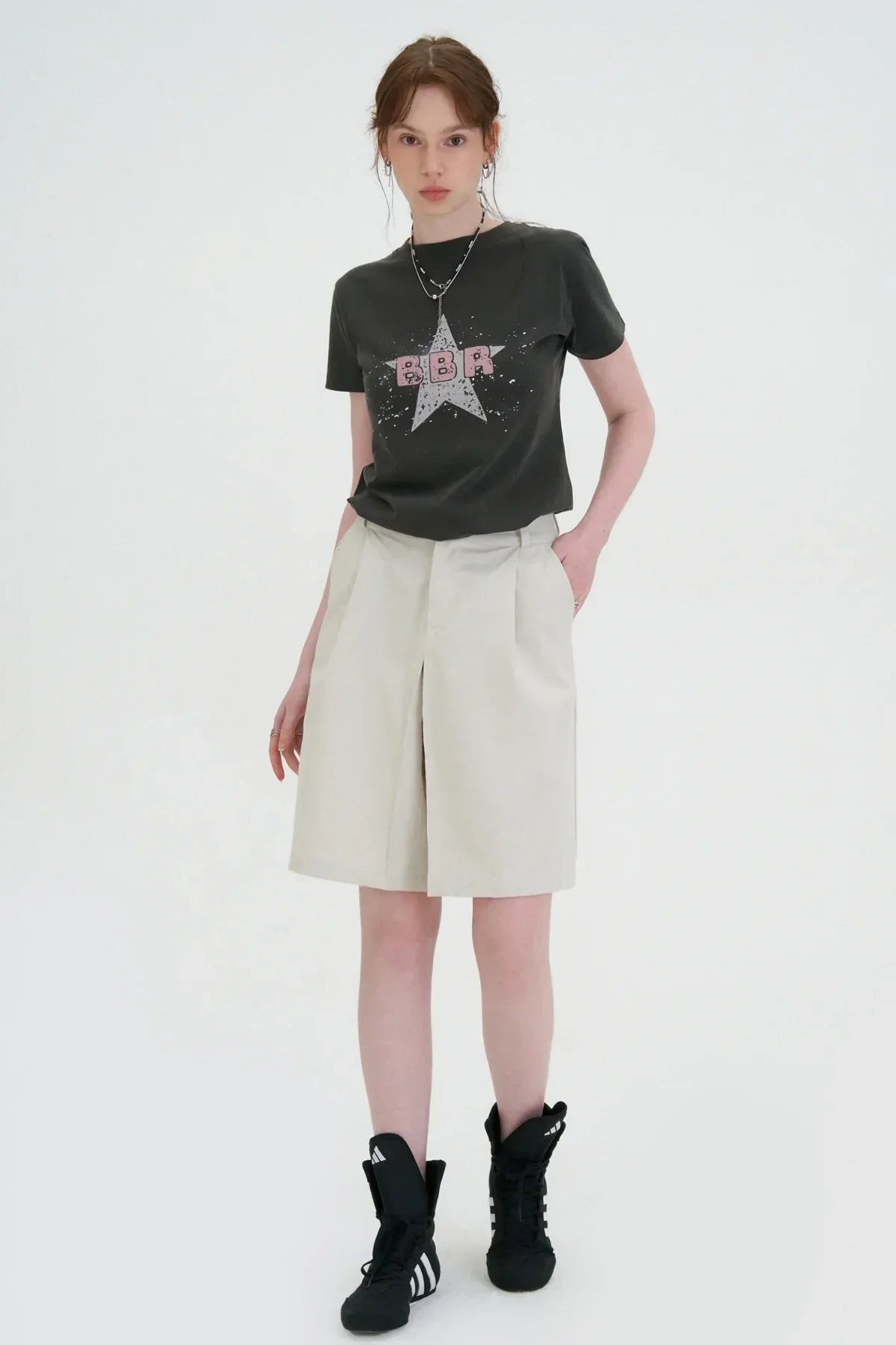 Young woman wearing charcoal graphic t-shirt, beige knee-length shorts, and black Adidas sneakers in studio
