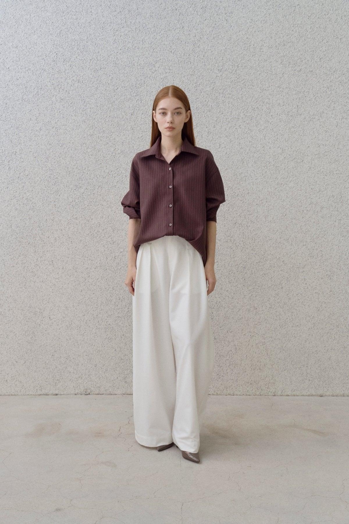 Woman in brown pinstripe shirt and wide-leg white pants standing against textured gray wall