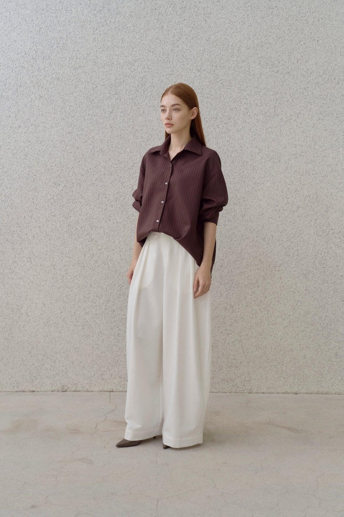 Young woman wearing a brown pinstripe shirt and wide white pants standing against a textured beige wall