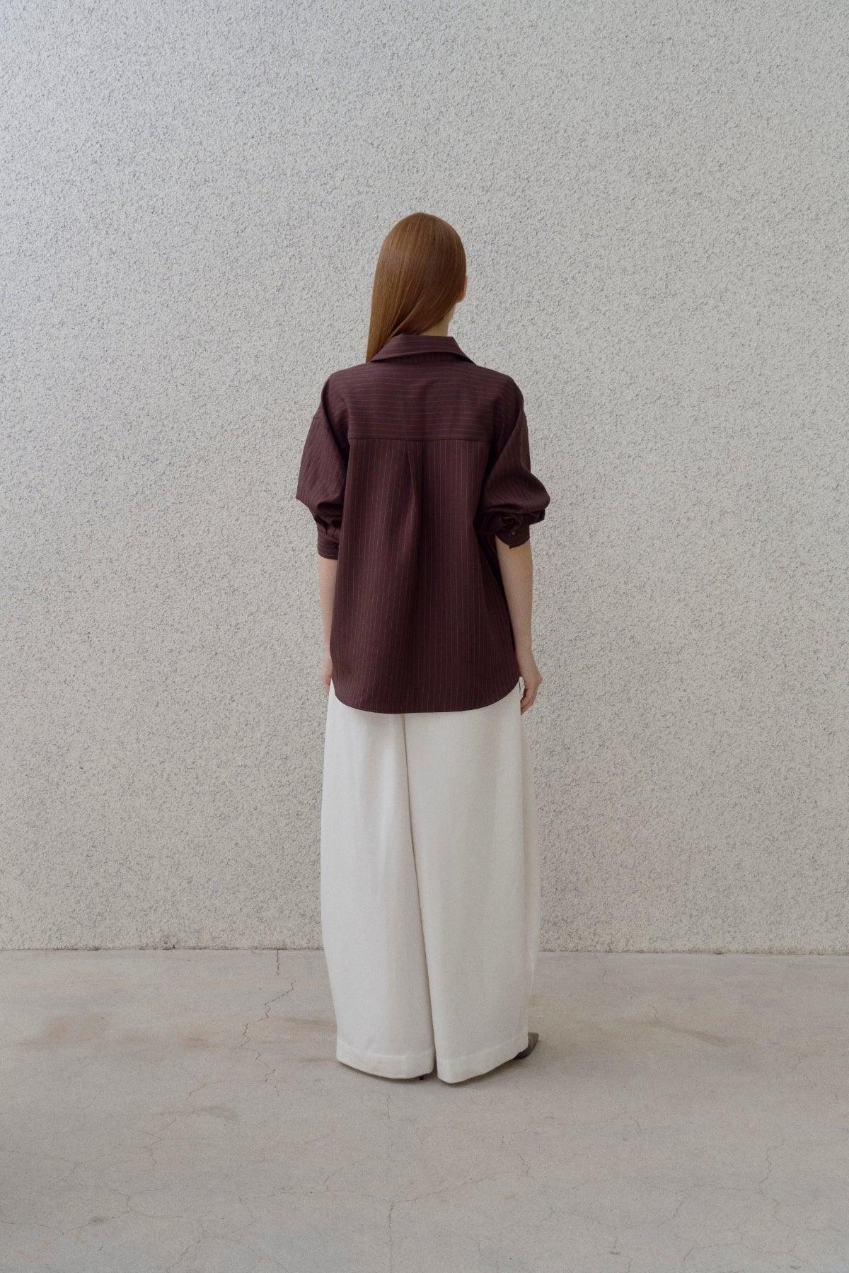 Woman wearing brown pinstripe shirt and white wide-leg pants standing against textured gray wall