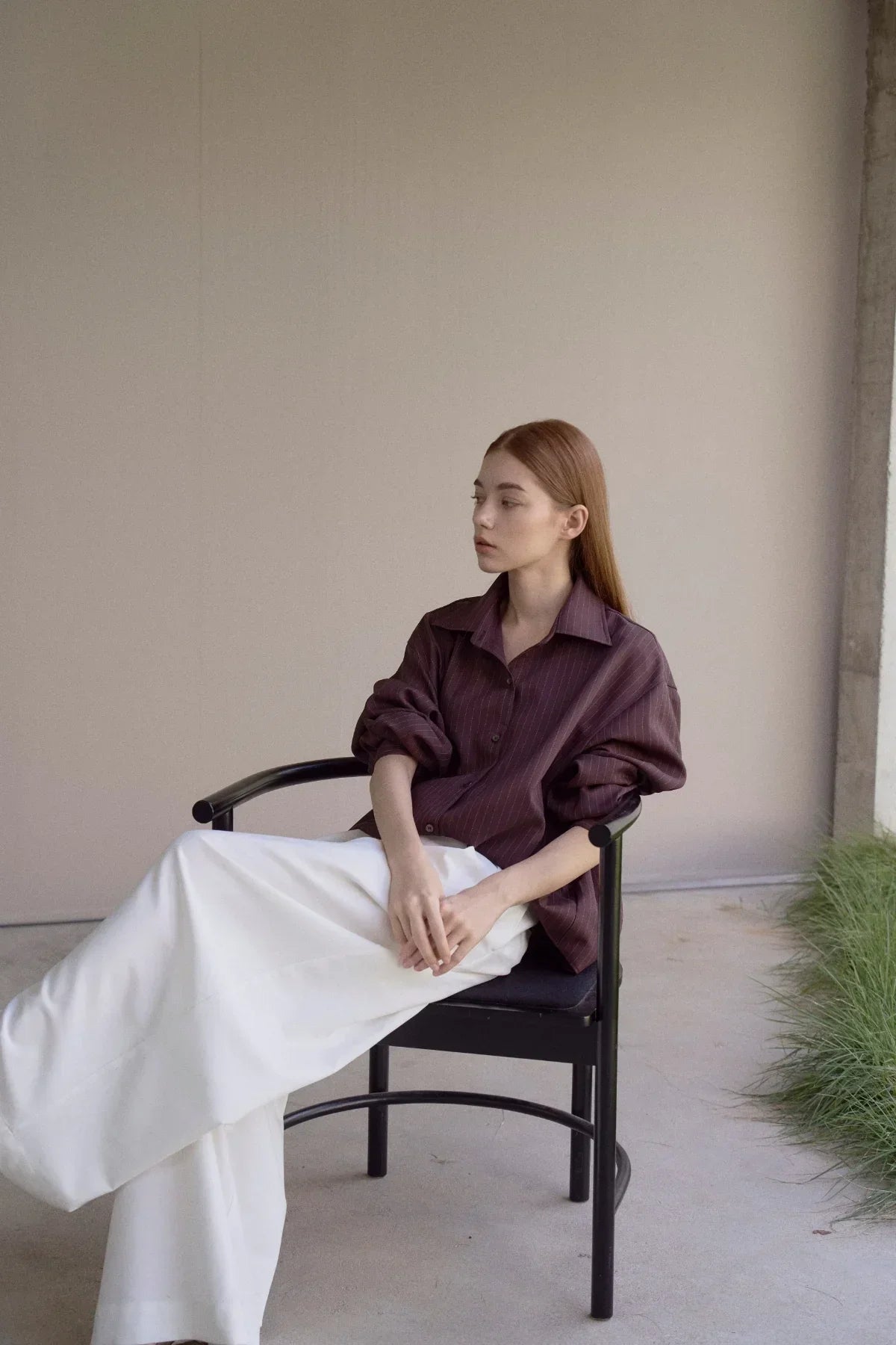 Woman in brown pinstripe shirt and white wide-leg pants sitting on black chair in minimalist space