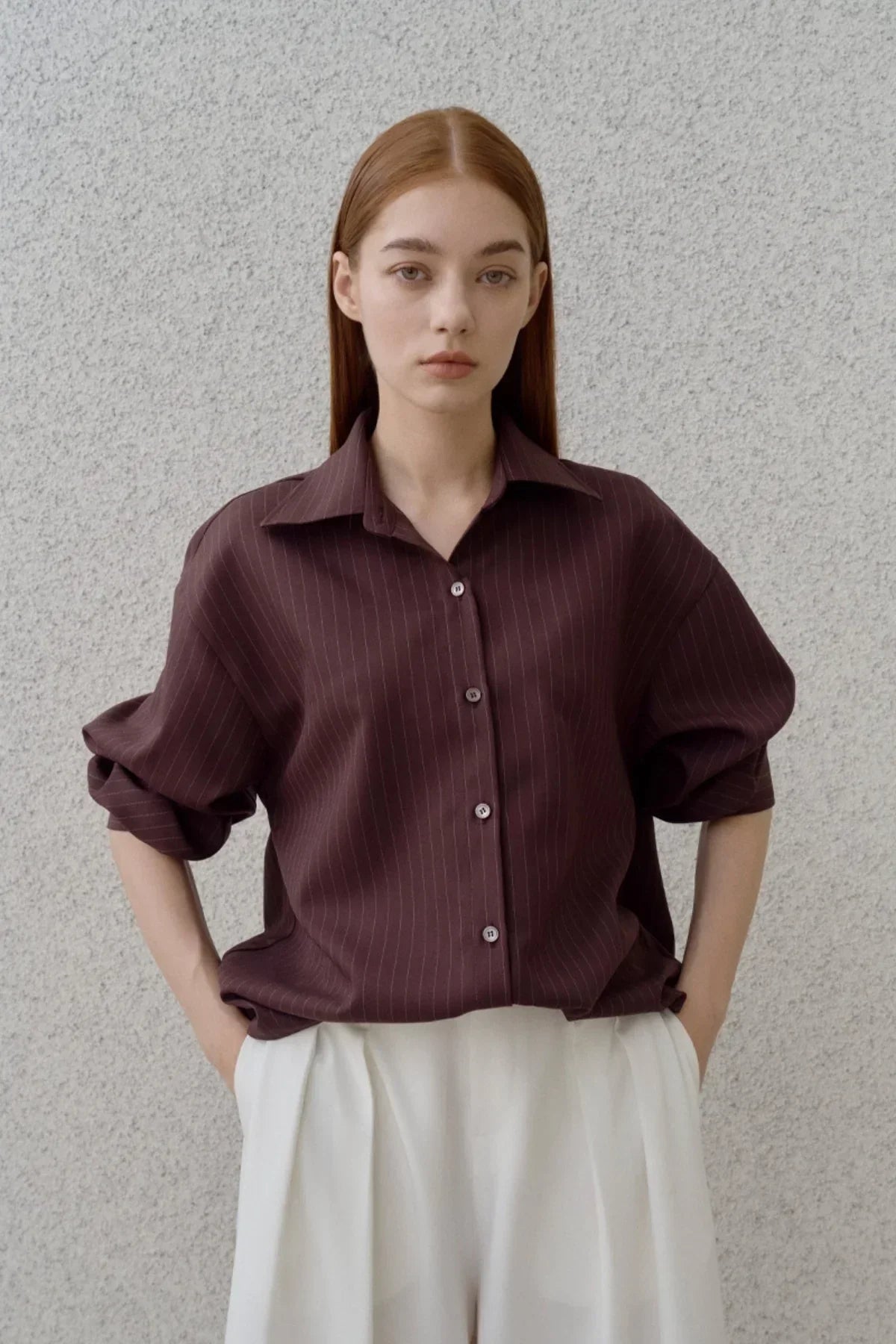 Young woman wearing a brown pinstripe oversized shirt with rolled-up sleeves and white pleated pants against a textured gray wall