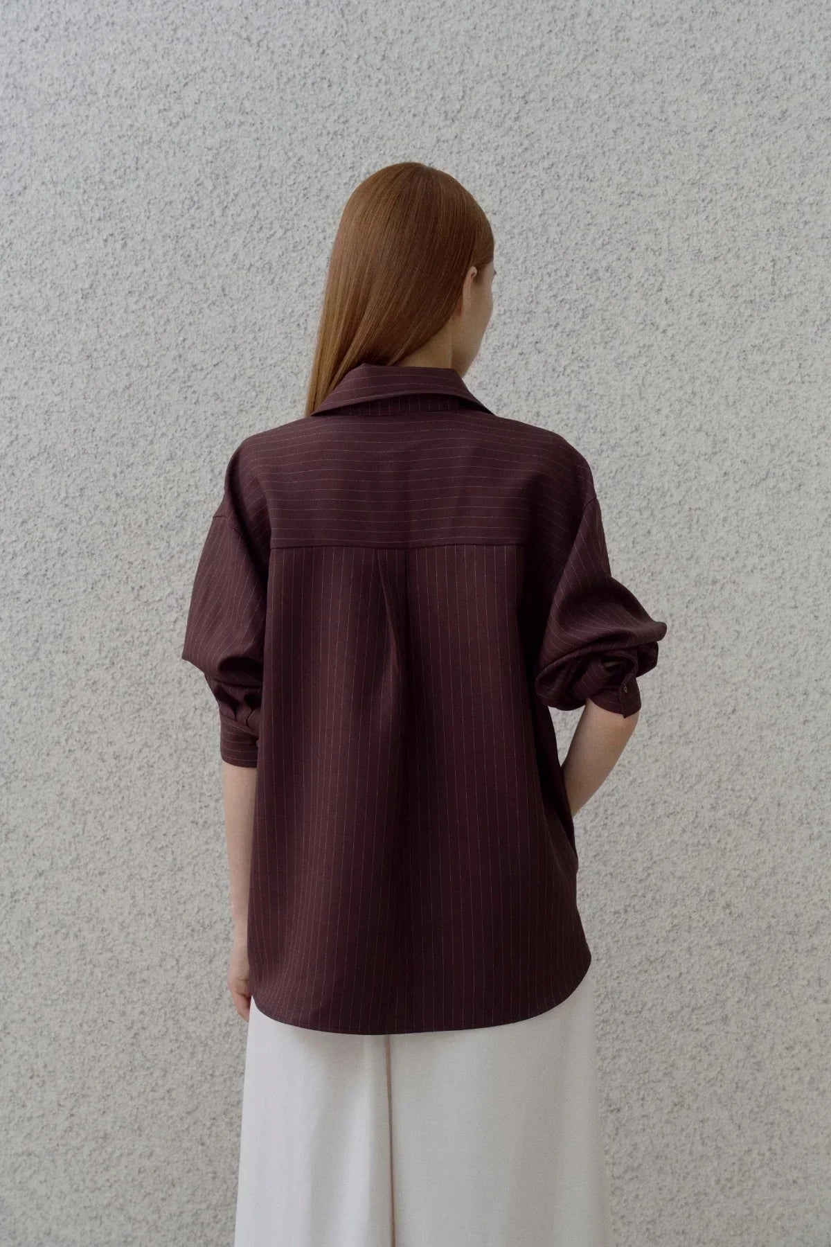 Back view of a woman wearing a brown pinstripe shirt with rolled-up sleeves against a textured wall
