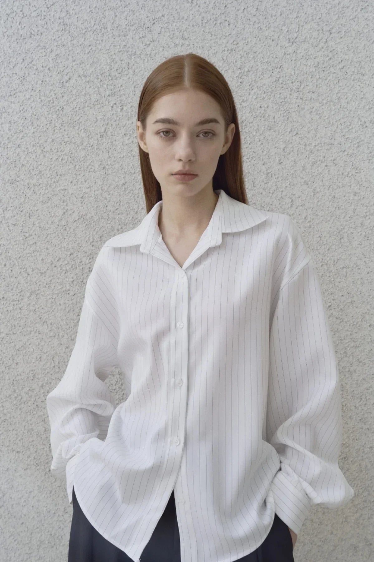 Young woman wearing oversized white pinstripe shirt posing against textured gray wall