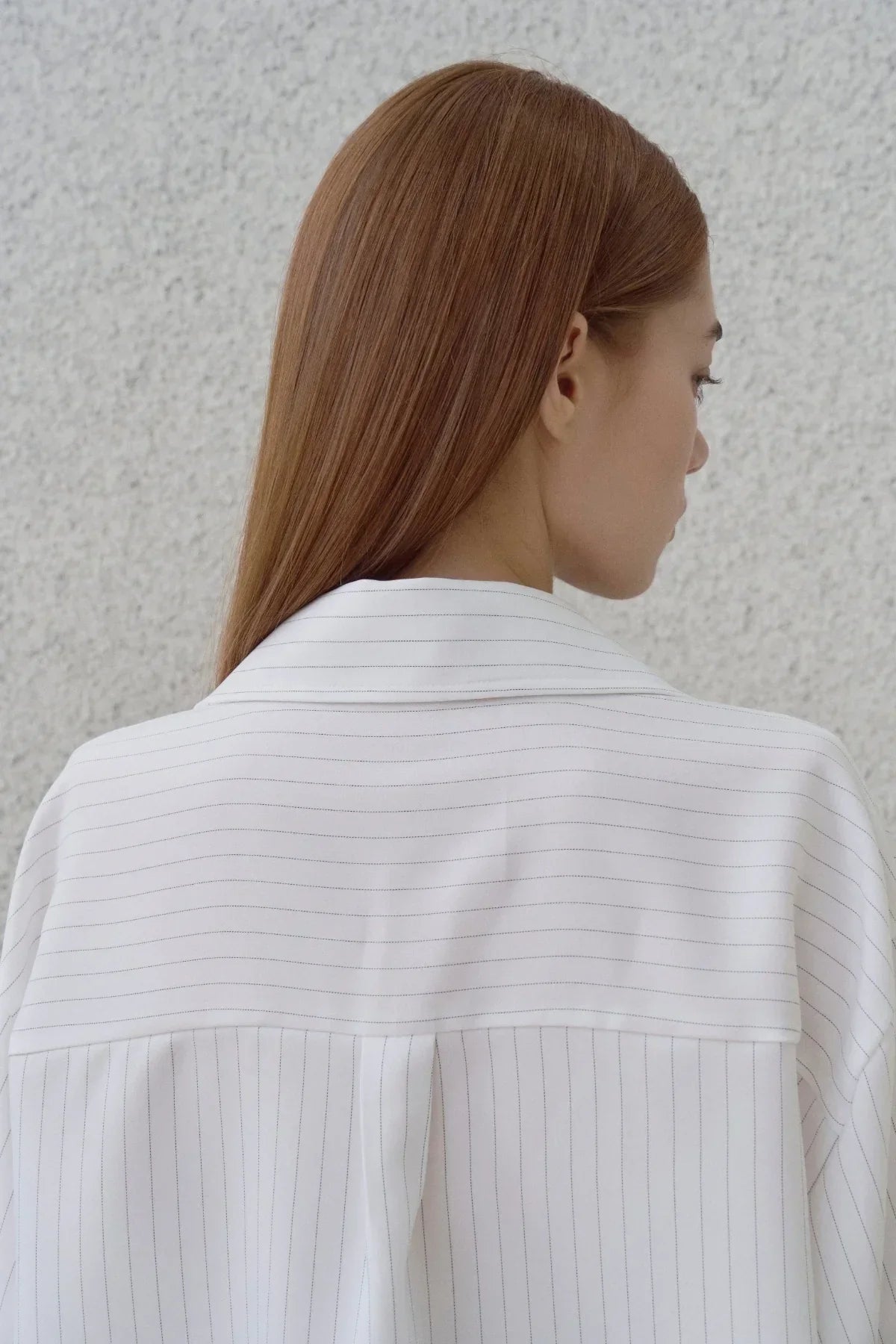 Back view of woman with straight brown hair wearing a white pinstripe shirt against gray wall