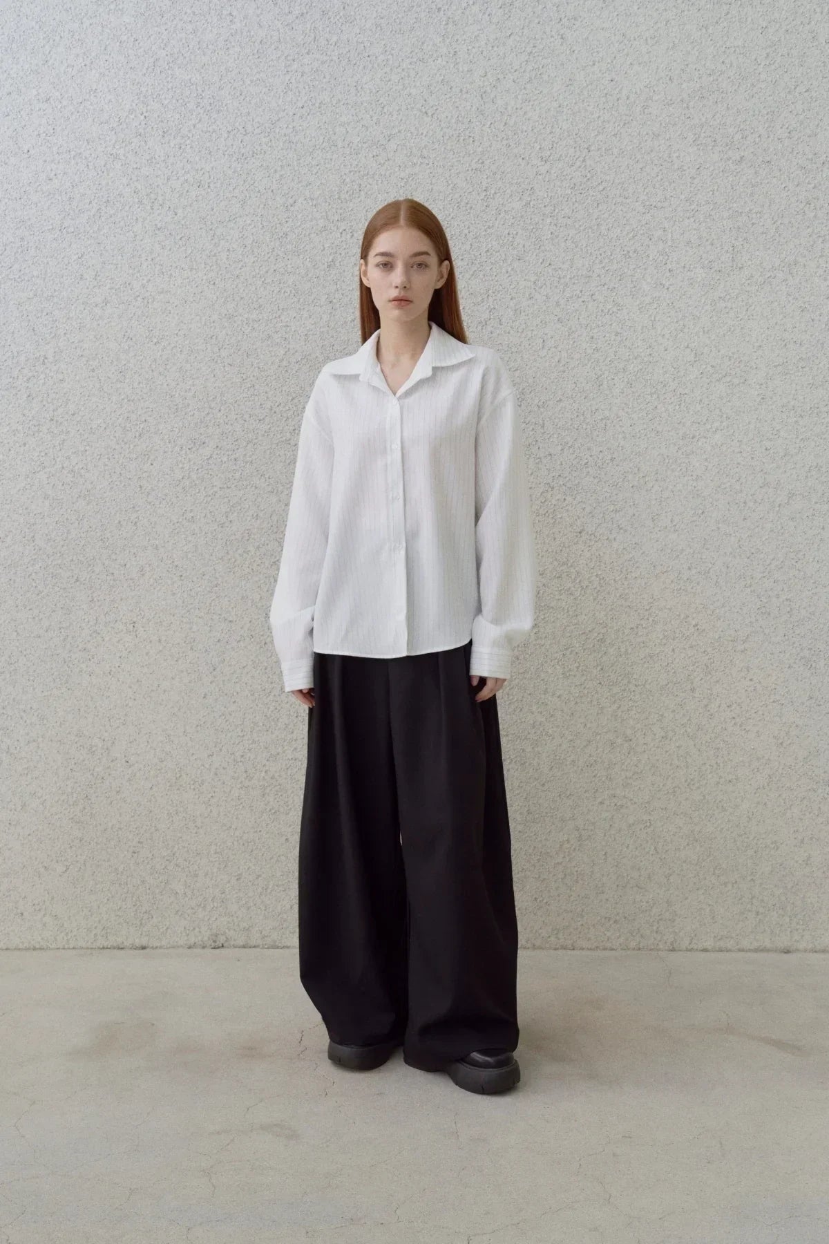 Woman wearing white pinstripe button-up shirt and wide black pants standing against textured gray wall