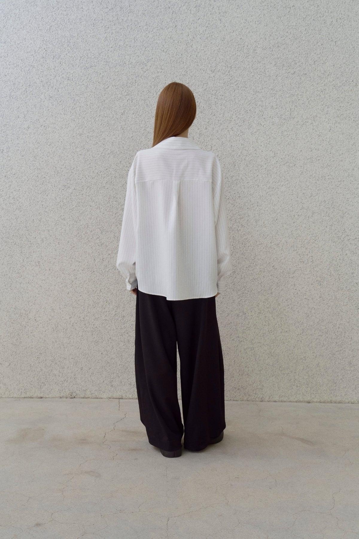 Back view of woman with straight hair wearing white pinstripe shirt and wide black pants against gray wall