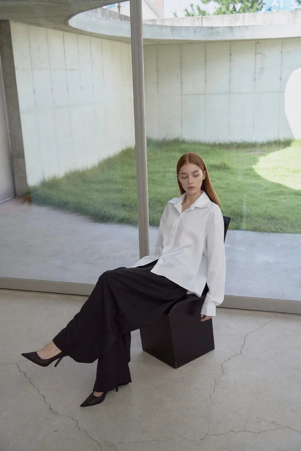 Woman in white pinstripe shirt and black wide-leg pants sitting on black chair in modern minimal room