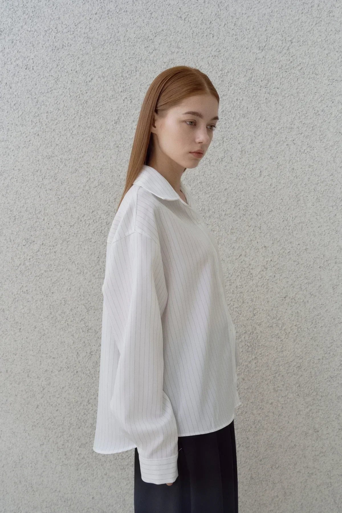 Side view of woman wearing oversized white pinstripe shirt and black pants against gray textured wall