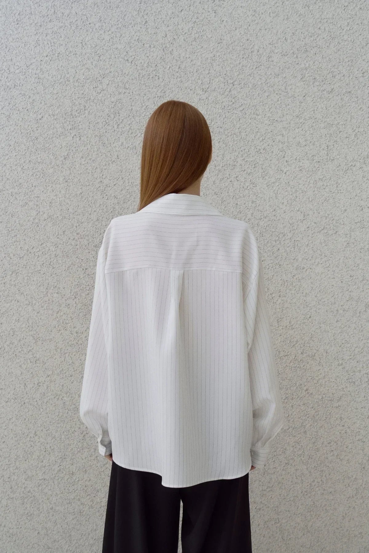 Back view of person with straight hair wearing white pinstripe shirt and black pants against textured wall