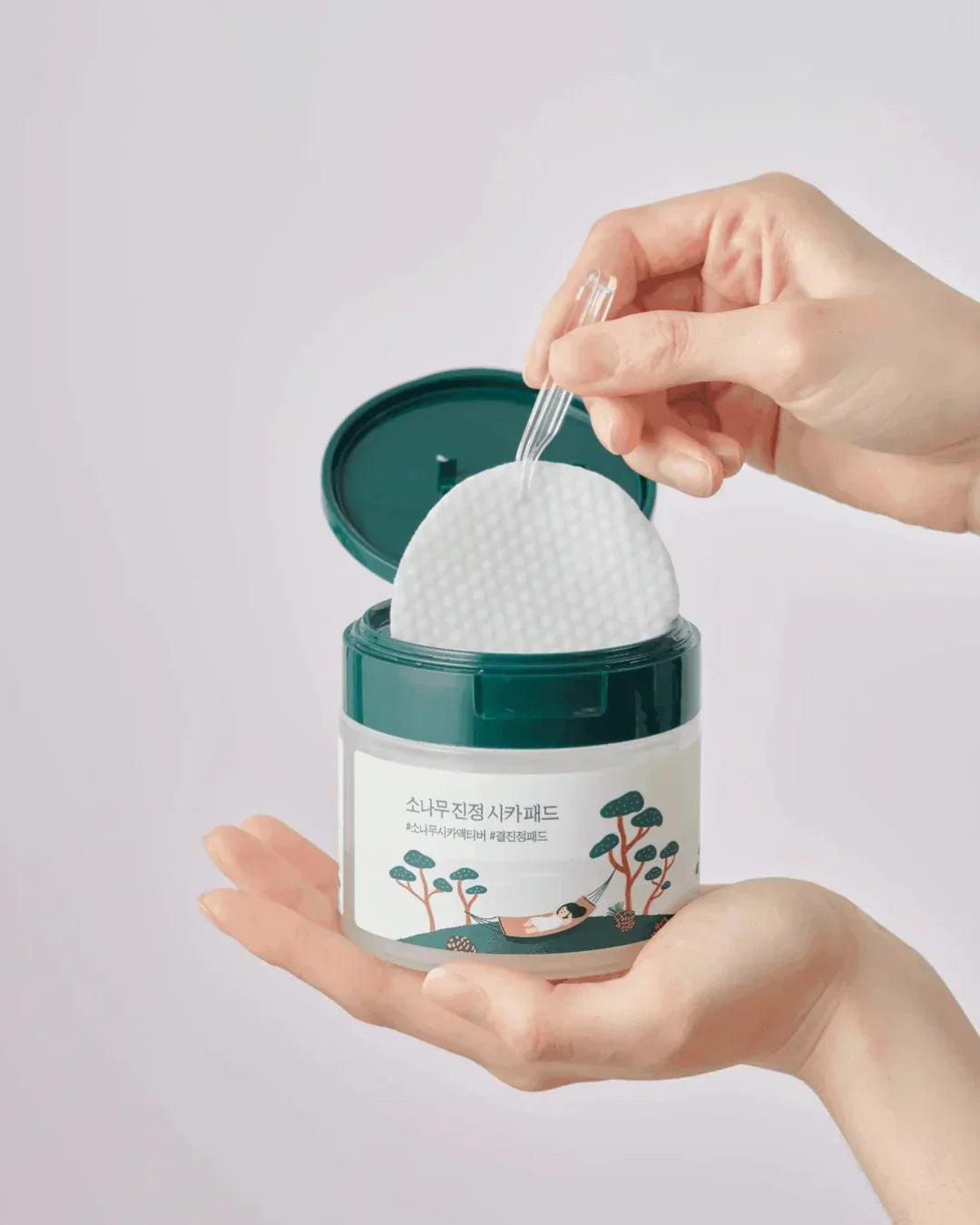 Hand holding open green container of pine calming cica pads with another hand using tweezers to lift a pad