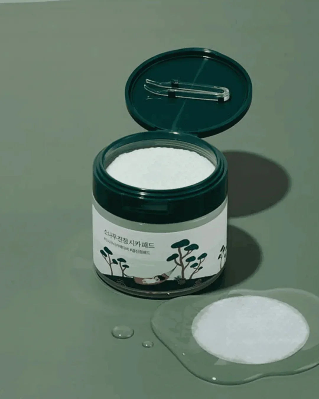 Open jar of Pine Calming Cica Pads with white cotton pads and tweezers on green background