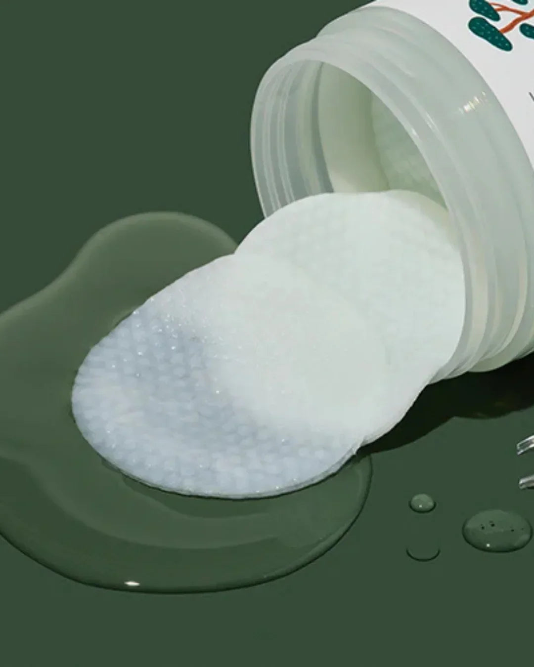 White textured calming cica facial pads spilling from container on dark green surface with liquid droplets