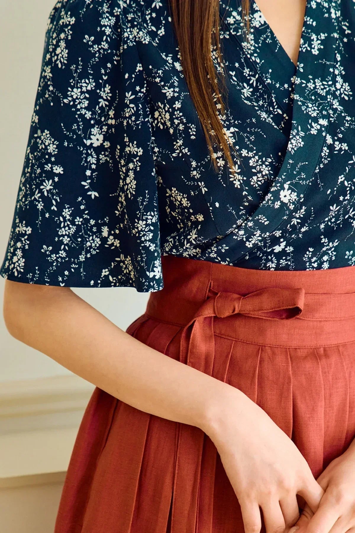 Close-up of woman wearing navy floral blouse with flutter sleeves and high-waisted rust pleated skirt with bow