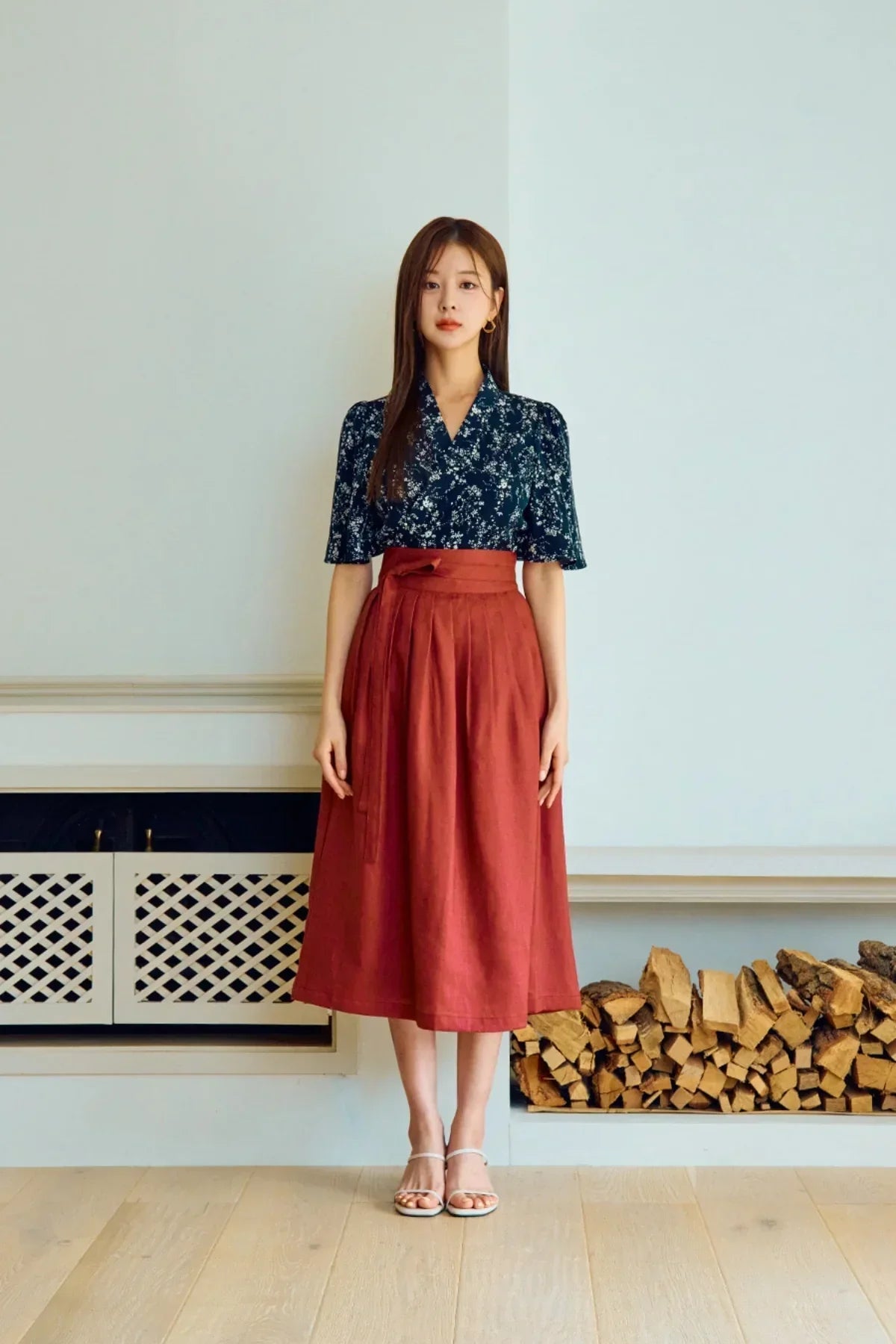 Woman wearing floral navy blouse and rust red midi skirt standing indoors by stacked firewood