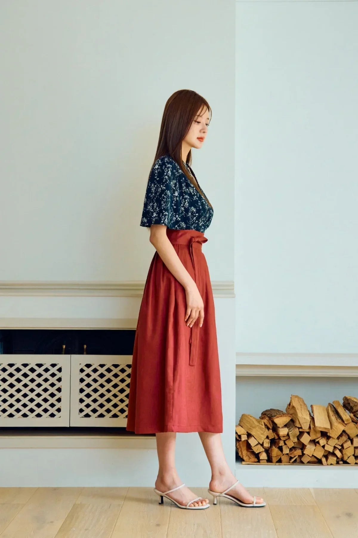 Woman wearing a navy floral blouse and rust-colored high-waist midi skirt indoors by stacked firewood