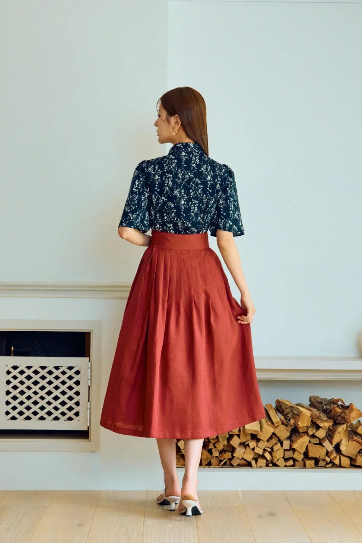 Woman wearing navy floral blouse and high-waisted rust midi skirt standing indoors near stacked firewood