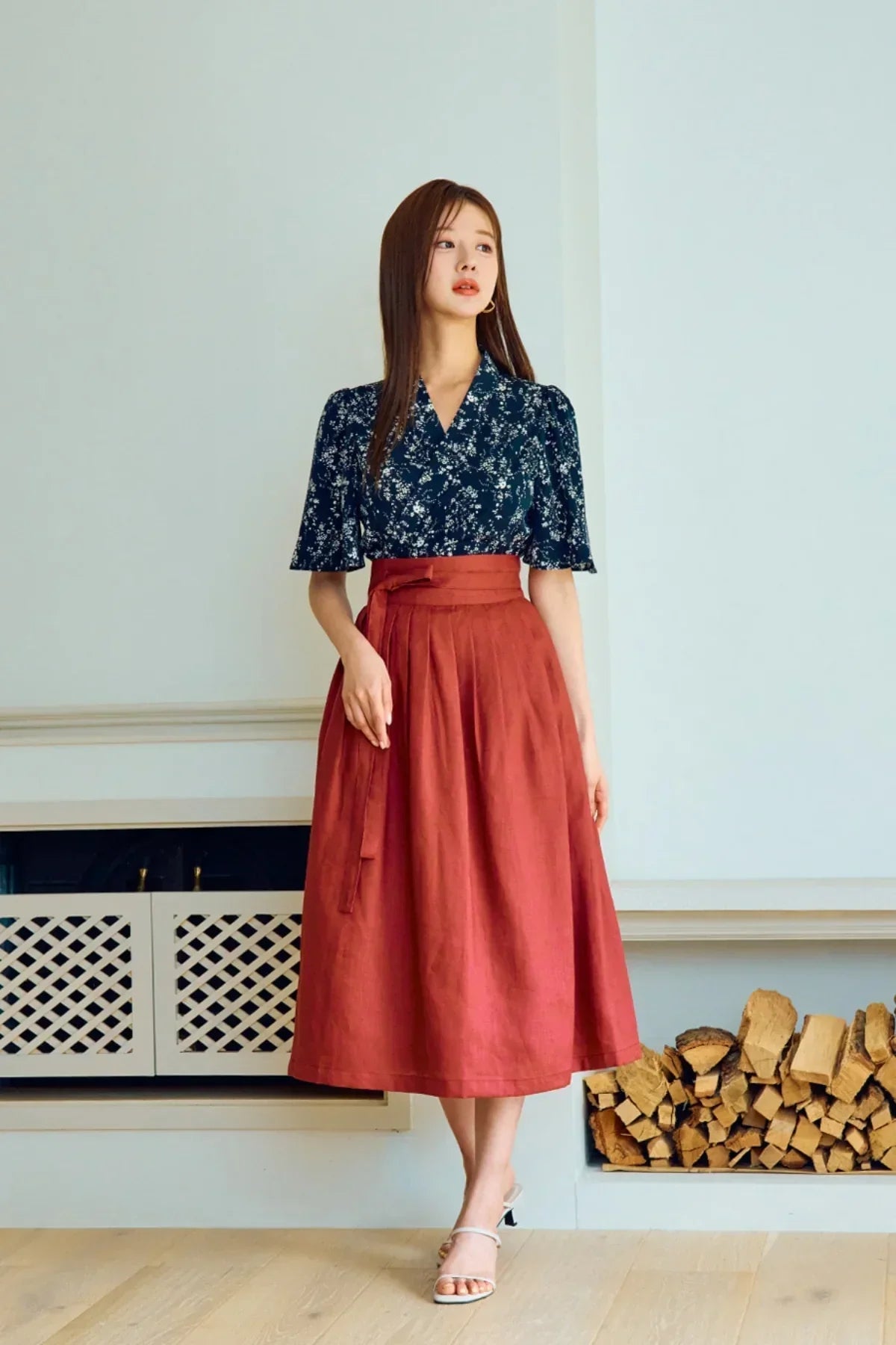 Woman wearing navy floral blouse and rust high-waisted midi skirt, standing indoors near wood stack