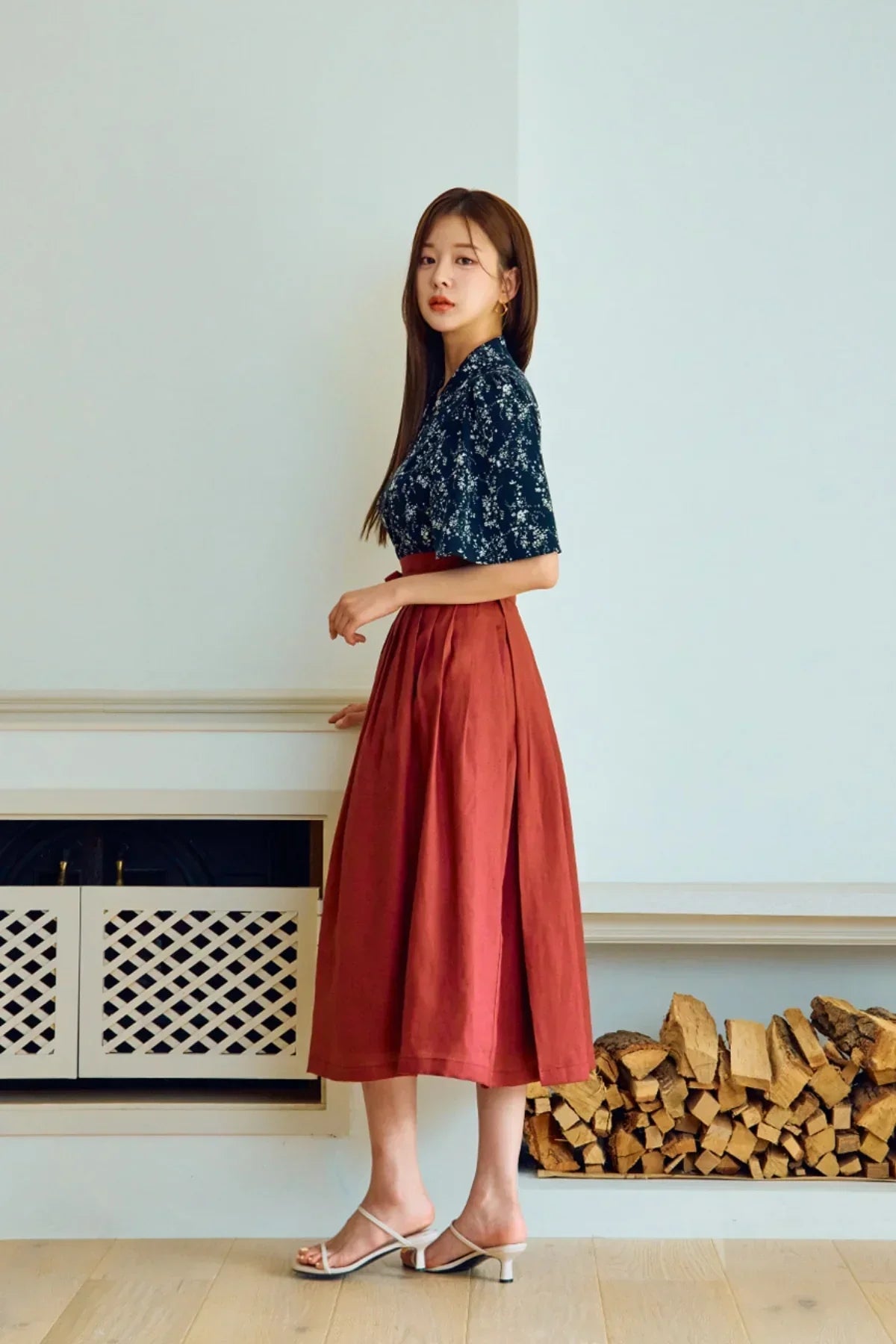 Woman wearing a navy floral blouse with a rust-red midi skirt and white heeled sandals indoors