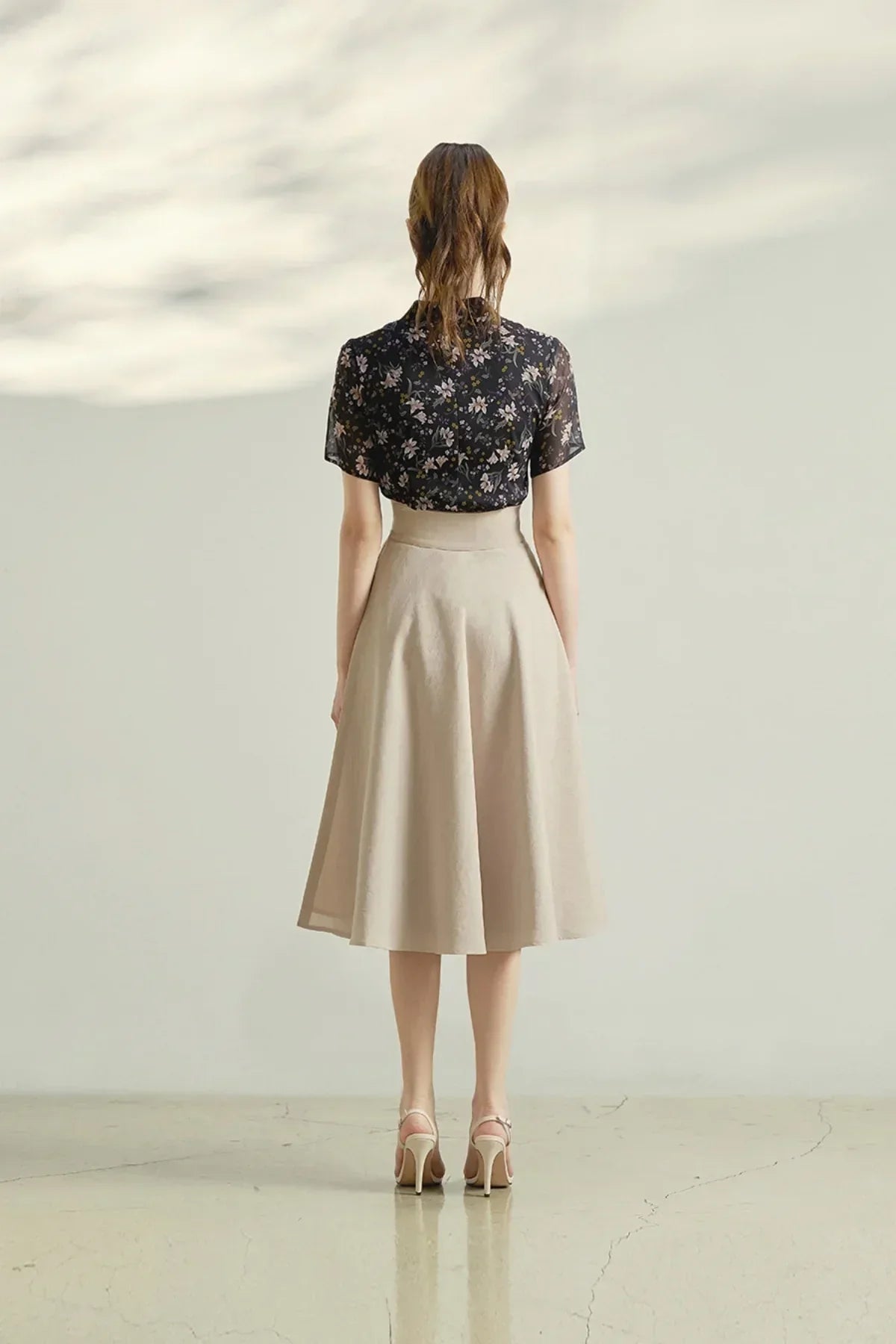 Woman wearing beige midi skirt and black floral blouse standing on polished floor with shadowed wall