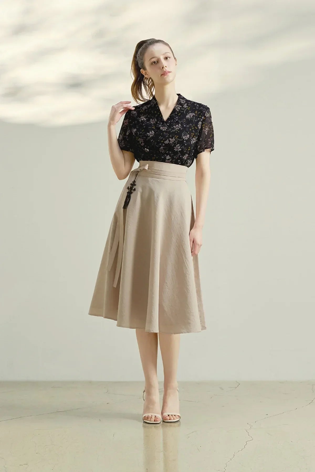 Woman wearing a black floral blouse and beige midi pinwheel skirt in minimalist studio