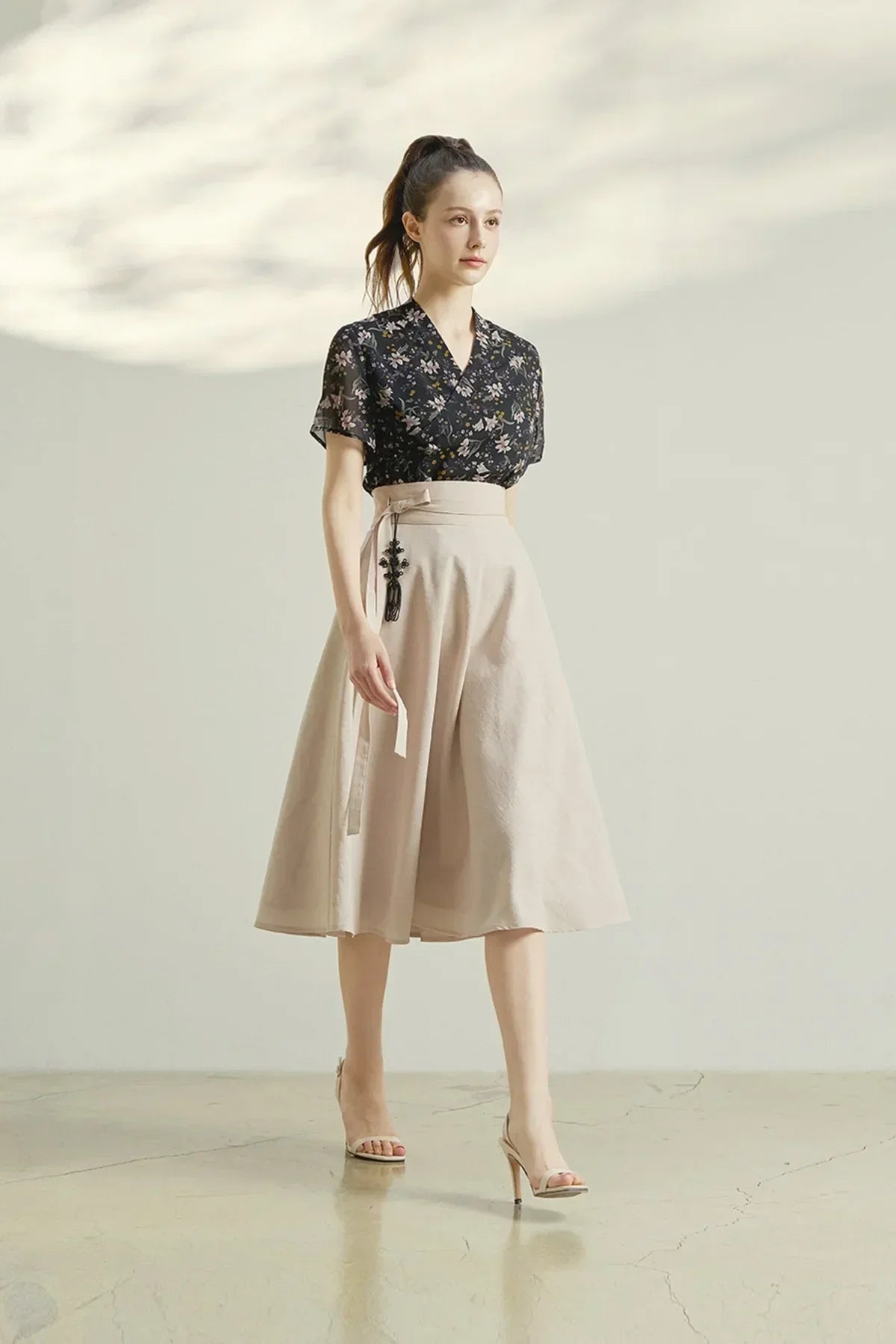 Woman in black floral blouse and beige midi pinwheel skirt walking on polished floor in studio