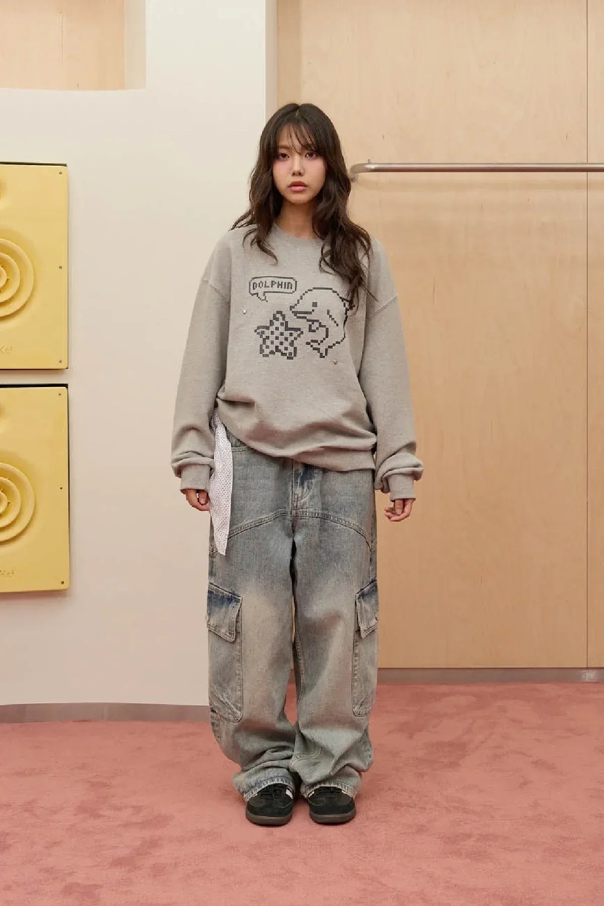 Woman wearing grey sweatshirt with pixel dolphin and star design, baggy jeans, black shoes, indoor setting